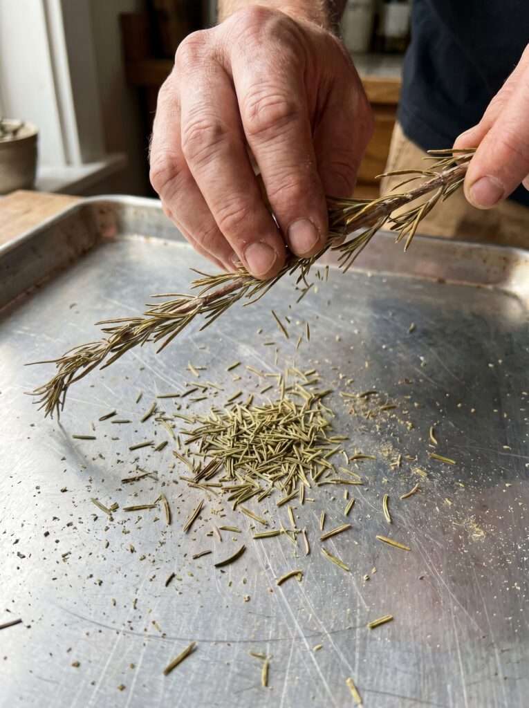 Hands stripping dried rosemary needles from the stem over a metal baking sheet.