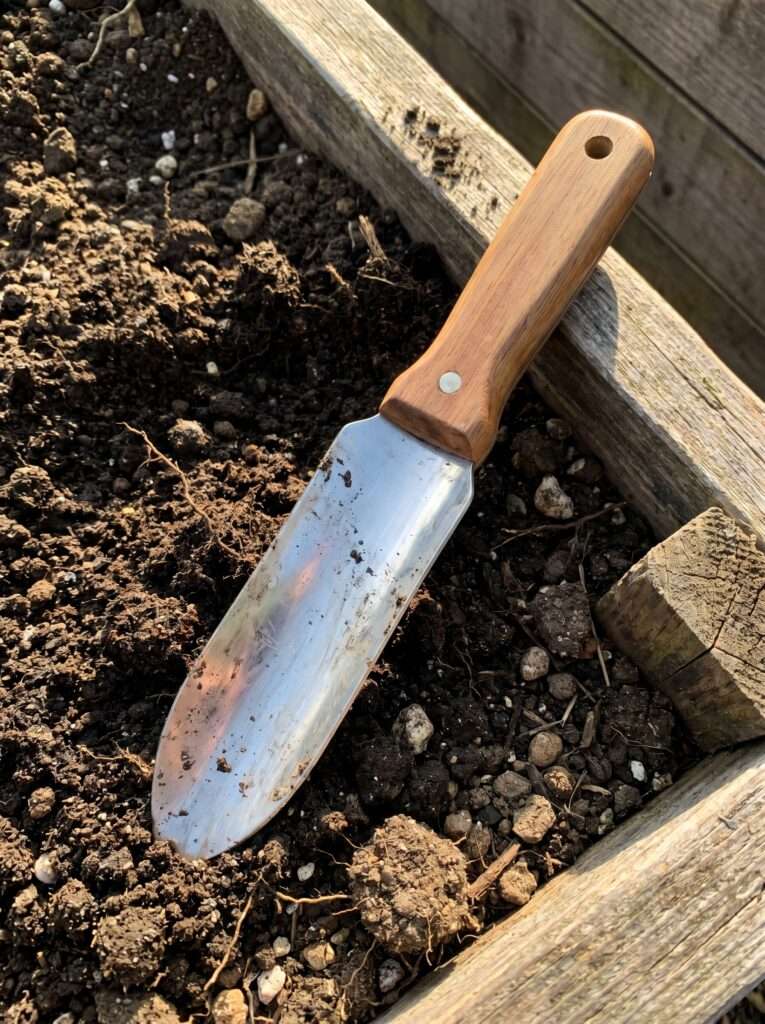 Worn Japanese Hori Hori gardening knife resting on dark soil in a raised bed