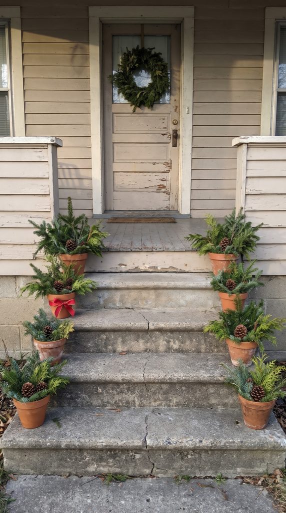 Small evergreen pots decorated with pinecones and red bows beside the steps of a Christmas-decorated front porch.