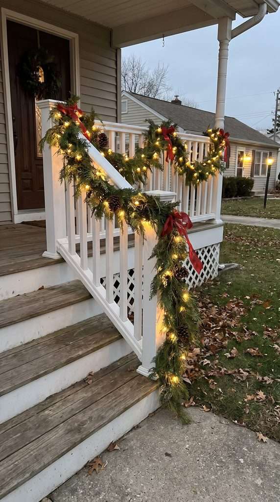 Evergreen Christmas garland with warm white lights and red bows wrapped around an American home's front porch railing.