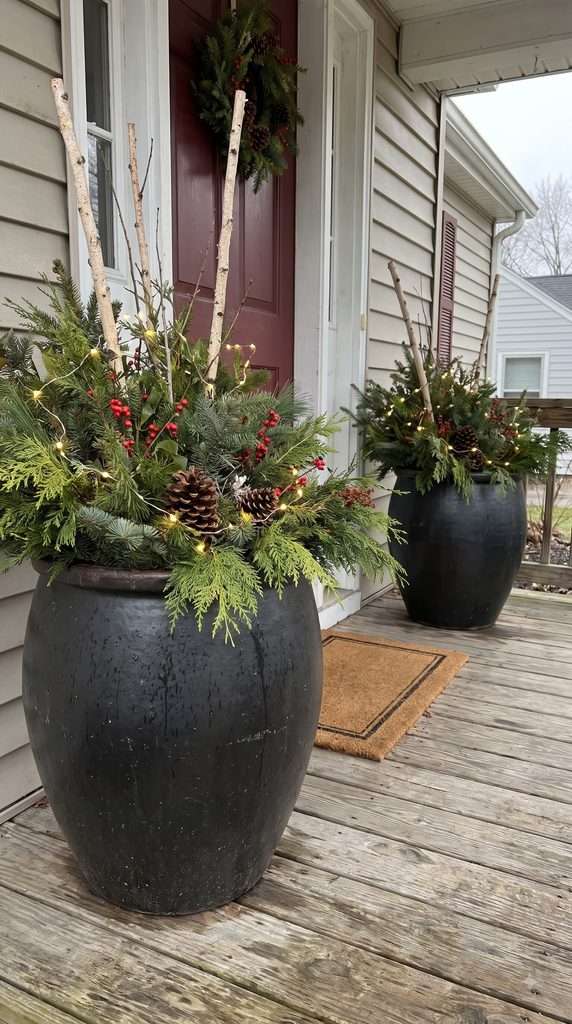 Large outdoor planters filled with evergreen branches, pinecones, red berries, and warm mini lights beside a Christmas front entrance.