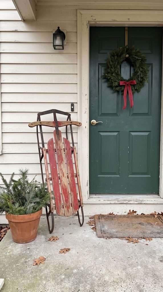 Worn wooden sled decorated with evergreen branches near a Christmas entryway.