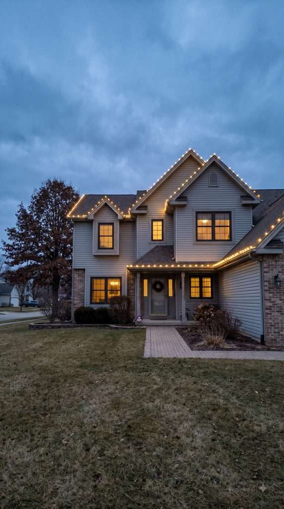 American suburban house with warm white Christmas lights outlining the roofline and front porch at dusk.